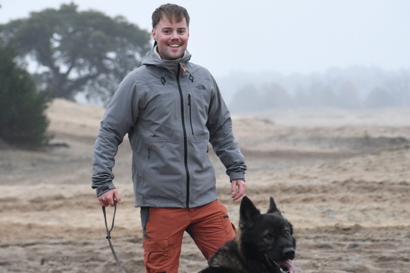 Man in grijze jas met een hond aan de lijn op een mistige, open locatie.